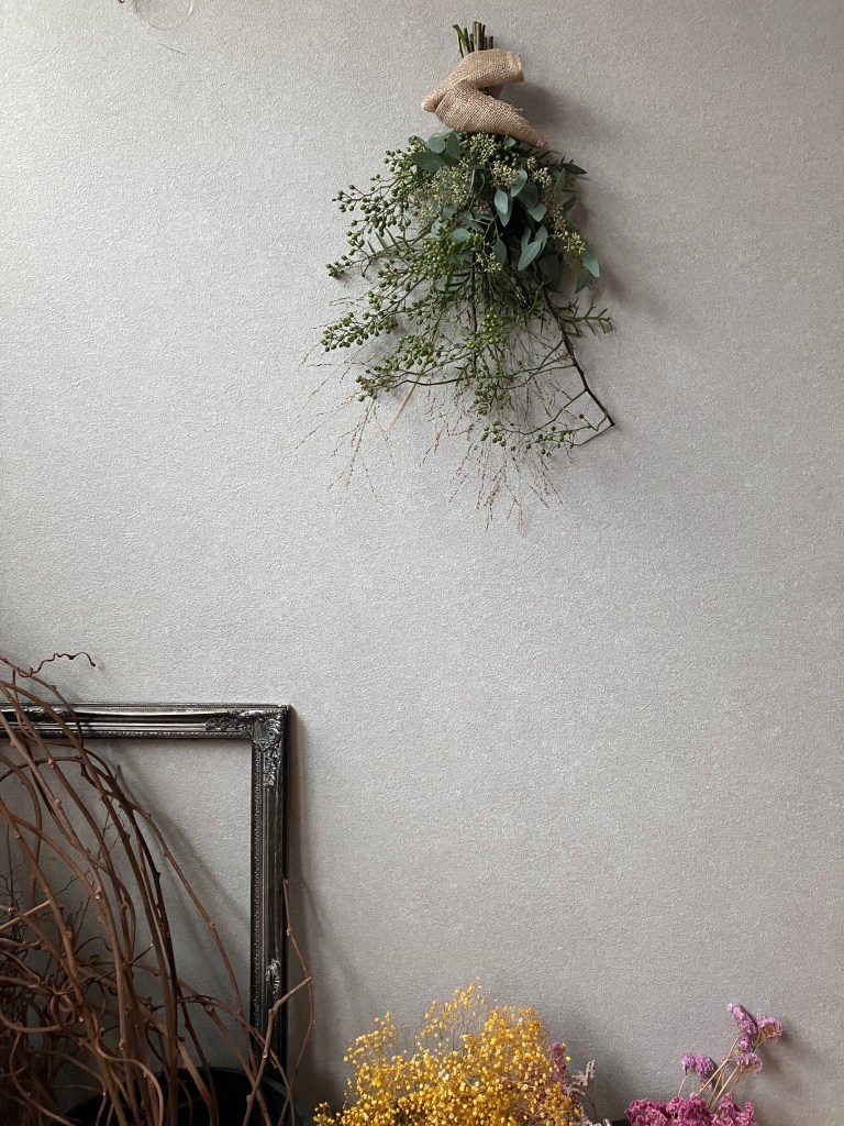 グレーの壁に飾られたフレッシュグリーンのスワッグ。足元にはドライフラワーが置いてある。少し遠くから全体を映している。