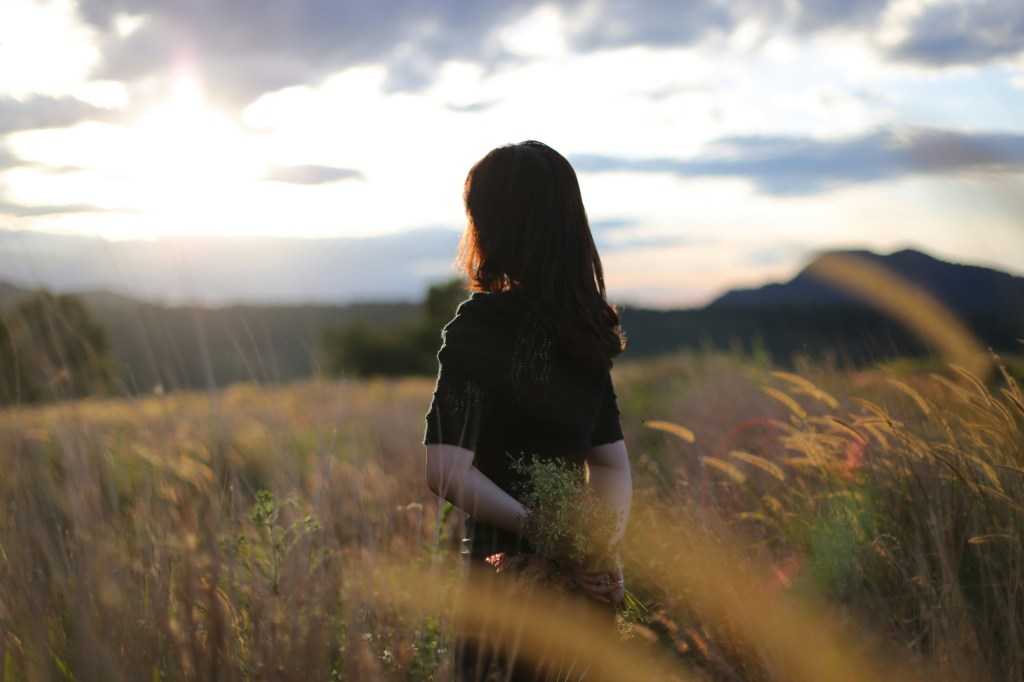 夕暮れの草原で、手に小さな花束を持ち、遠くを見つめる女性